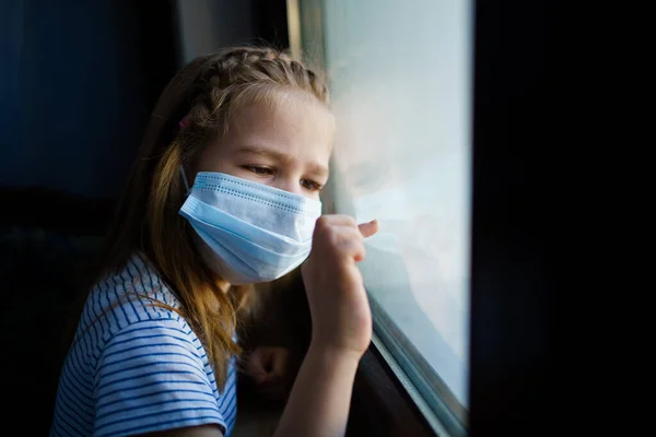 Niña con máscara protectora, mira por la ventana de afuera. autoaislante. — Foto de Stock