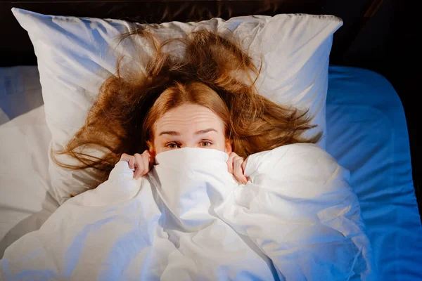 Vista dall'alto. una donna giace a letto sotto una coperta bianca e spaventata, spaventata. — Foto Stock