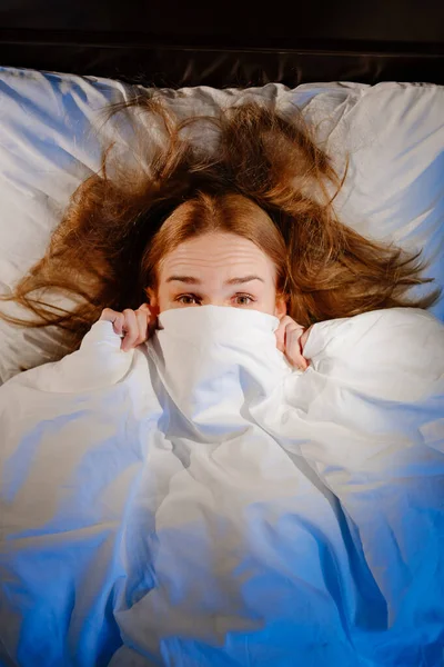 Vista dall'alto. una donna giace a letto sotto una coperta bianca e spaventata, spaventata. — Foto Stock