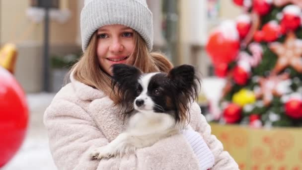 Kışın Noel sokaklarında kollarında Papillon köpeği olan güzel bir kız. — Stok video