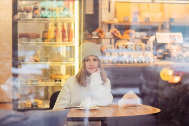 Şapkalı güzel bir kız camların arkasındaki şekerleme kafesinde oturuyor.