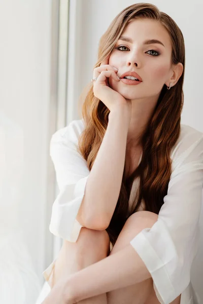 Hermosa mujer en túnica blanca se sienta en windowsill.morning de la novia antes de la boda —  Fotos de Stock