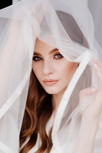 Porträtt av en vacker ung kvinna i en vit morgonrock under en bröllopsslöja. — Stockfoto