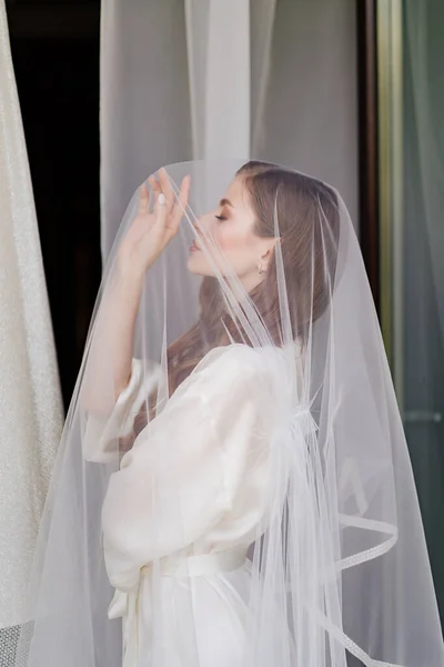 Portret van een mooie jonge vrouw in een wit gewaad onder een trouwsluier. — Stockfoto