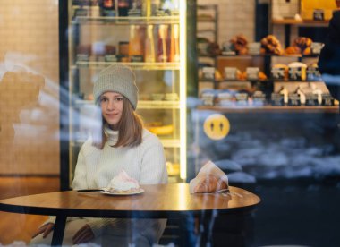 Şapkalı güzel bir kız camların arkasındaki şekerleme kafesinde oturuyor.