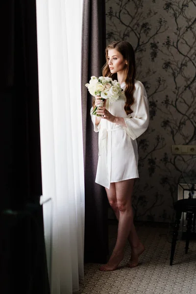 Mooie vrouw in een witte badjas met een bruiloft boeket van bloemen aan het venster — Stockfoto