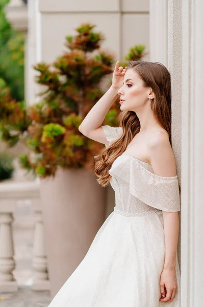 Schöne Frau mit langen Haaren im Brautkleid steht an Hauswand. — Stockfoto