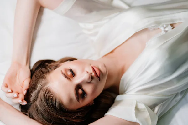 Θέα από πάνω. σέξι γυναίκα με μακιγιάζ, μακριά μαλλιά βρίσκεται σε λευκό ρόμπα στο κρεβάτι — Φωτογραφία Αρχείου