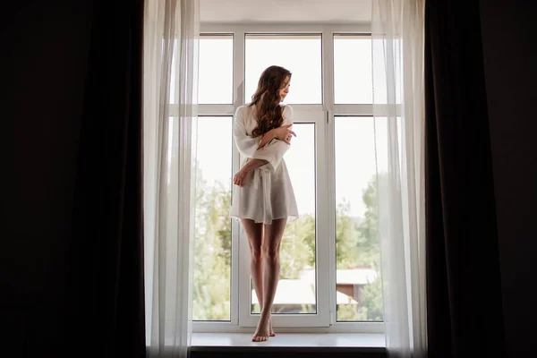 Beautiful sexy girl in a white robe with long hair stands in the window opening