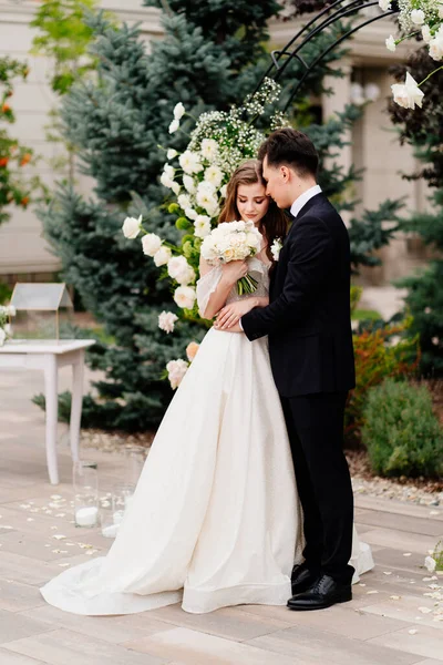 Ceremonia de boda en arco de flores vivas. Hermosos recién casados abrazan tiernamente —  Fotos de Stock