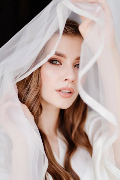 Retrato de uma bela jovem mulher em um manto branco sob um véu de casamento. — Fotografia de Stock