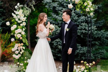 Açık havada, canlı çiçeklerin kemerinde bir düğün töreni. Gelin duygulanır ve ağlar.