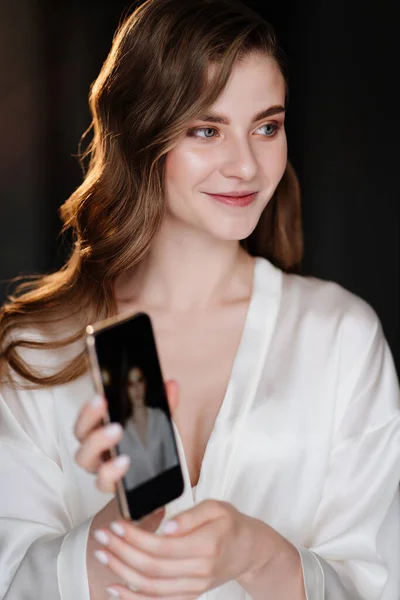 The beautiful woman looks at her photo on the phone with makeup and styling.