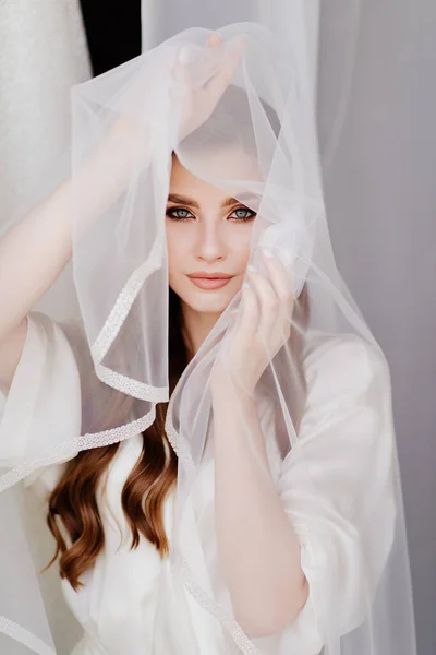 Retrato de uma bela jovem mulher em um manto branco sob um véu de casamento. — Fotografia de Stock