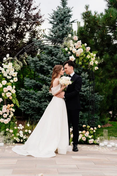 Hochzeitszeremonie im Bogen lebender Blumen. Schöne Brautleute umarmen sich zärtlich — Stockfoto