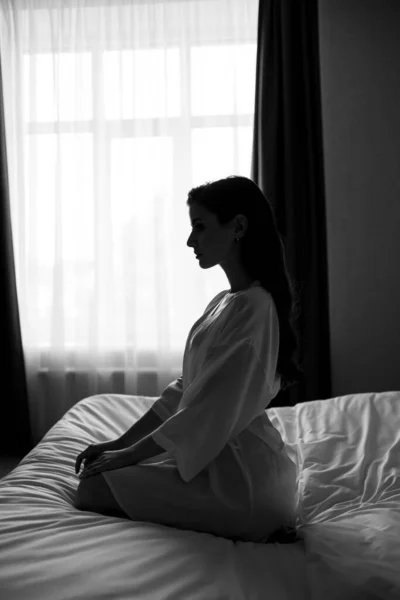 Silhouette. beautiful woman with makeup, long hair sits in white robe on bed — Stock Photo, Image