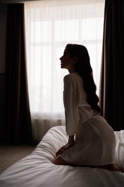 Silüet. Makyajlı güzel bir kadın, uzun saçlı beyaz bornozlu yatakta oturuyor.