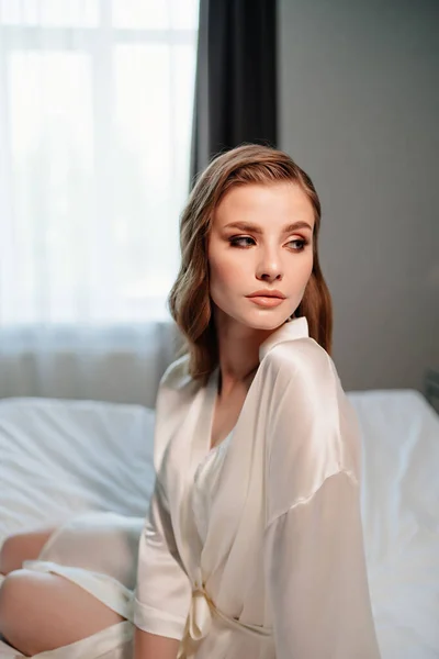Bela mulher com maquiagem, cabelos longos senta-se em roupão branco na cama — Fotografia de Stock