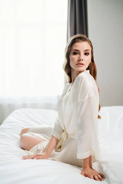 Hermosa mujer con maquillaje, pelo largo se sienta en bata blanca en la cama — Foto de Stock