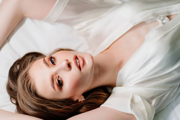 Vista desde arriba. sexy mujer con maquillaje, pelo largo se encuentra en túnica blanca en la cama —  Fotos de Stock