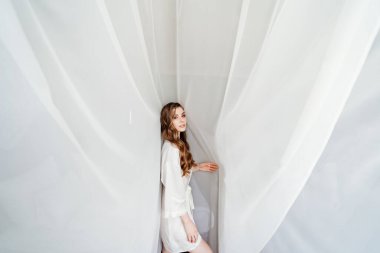 Uzun saçlı, beyaz cüppeli güzel bir kadın perdenin altında duruyor.