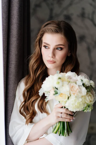 하얀 가운을 입고 결혼식 꽃다발을 창문에 걸고 있는 아름다운 여인 — 스톡 사진