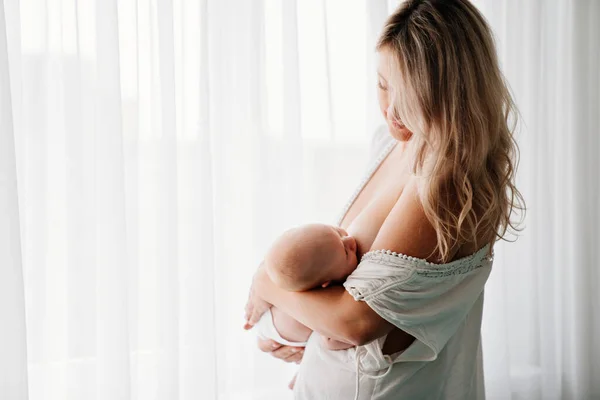 Mamá alimenta al bebé de pie en la ventana. amamantar. — Foto de Stock