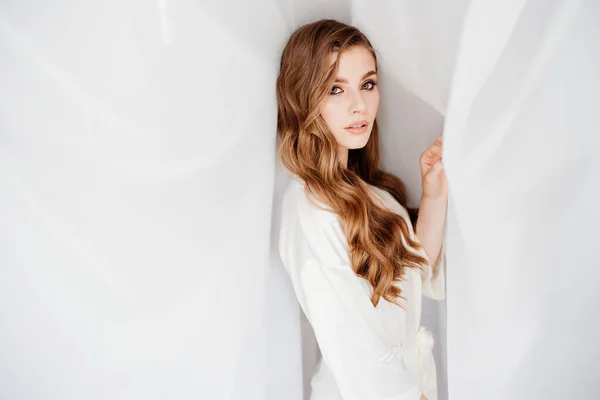 Hermosa joven con el pelo largo en una túnica blanca se encuentra debajo de una cortina — Foto de Stock