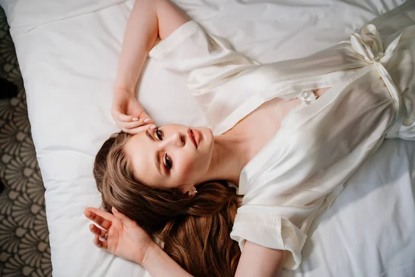 Vista desde arriba. sexy mujer con maquillaje, pelo largo se encuentra en túnica blanca en la cama —  Fotos de Stock