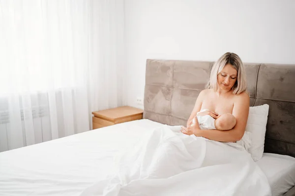 Sin ropa madre está sentada con un bebé en una cama blanca mamando. —  Fotos de Stock
