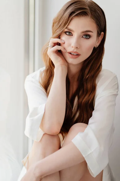 Hermosa mujer en túnica blanca se sienta en windowsill.morning de la novia antes de la boda — Foto de Stock