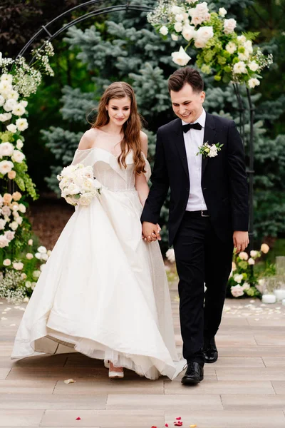 De beaux jeunes mariés vont à la cérémonie de mariage en plein air dans une arche de fleurs vivantes — Photo