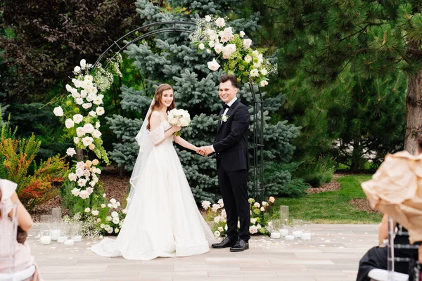 Utomhus bröllop ceremoni i en båge av levande blommor. Nygifta håller varandra i handen — Stockfoto