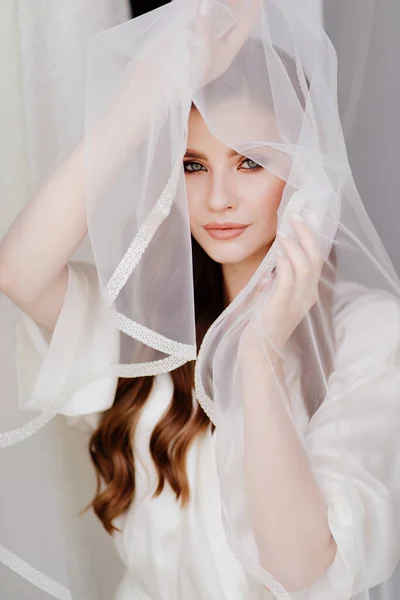 Retrato de uma bela jovem mulher em um manto branco sob um véu de casamento. — Fotografia de Stock