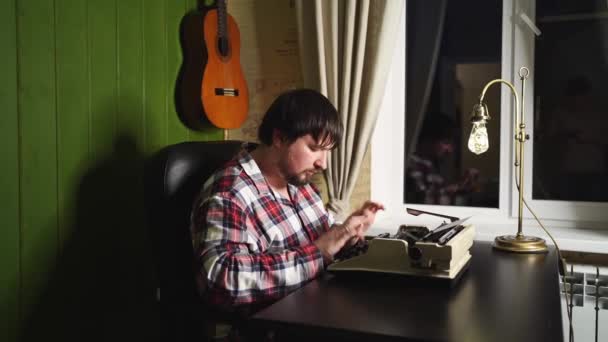 Ένας άντρας με καρό πιτζάμες δακτυλογραφεί σε μια γραφομηχανή τη νύχτα στο γραφείο του.. — Αρχείο Βίντεο