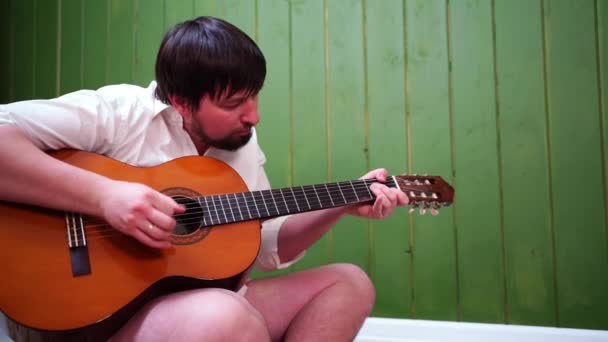 Um homem com uma barba em uma camiseta branca pela parede verde e toca guitarra. — Vídeo de Stock