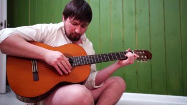 Yeşil duvarın yanında beyaz tişörtlü sakallı bir adam gitar çalıyor..