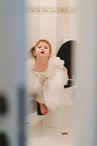 Una niña con un vestido elegante se sienta en el inodoro. disparando a través de las puertas. — Foto de Stock