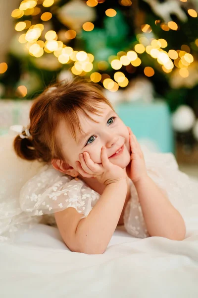Uma menina em um vestido branco jaz sob a árvore de Natal da casa. — Fotografia de Stock