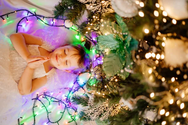 Menina com os olhos fechados em um vestido branco encontra-se sob a árvore de Natal da casa. — Fotografia de Stock