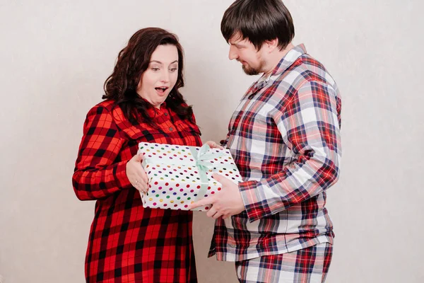 Ένα ζευγάρι σε κόκκινο καρό ρούχα στο σπίτι χέρια ένα δώρο γεμάτο με μια κορδέλα — Φωτογραφία Αρχείου