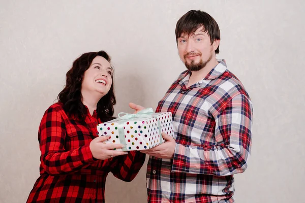 A couple in red checkered home clothes hands a gift packed with a bow of ribbon — Stock Photo, Image