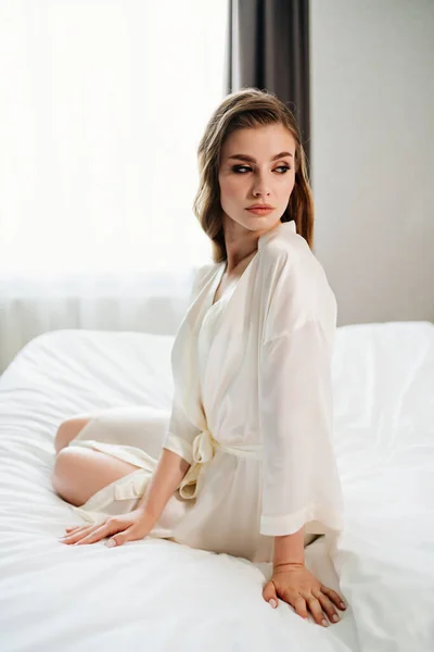 Hermosa mujer con maquillaje, pelo largo se sienta en bata blanca en la cama —  Fotos de Stock