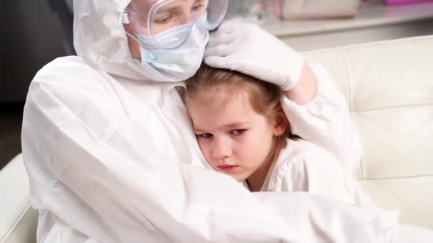 Доктор в защитном костюме, маске, перчатках и очках успокаивает маленькую больную девочку. — стоковое видео