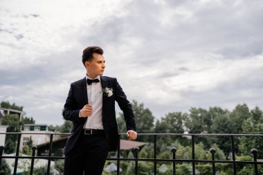 Siyah ceketli, elinde çiçek tomurcuğu olan genç bir adam balkonda duruyor. damat