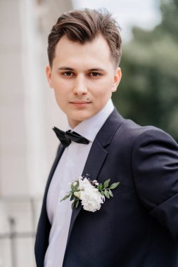 Siyah ceketli, çiçek tomurcuklu genç adam binanın yanındaki sokakta duruyor.