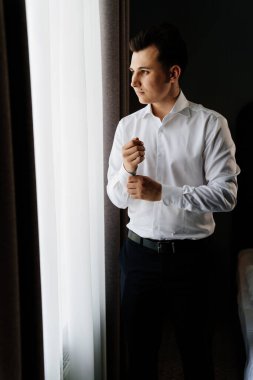 Beyaz gömlekli genç adam pencerenin yanındaki kol düğmelerini takıyor.