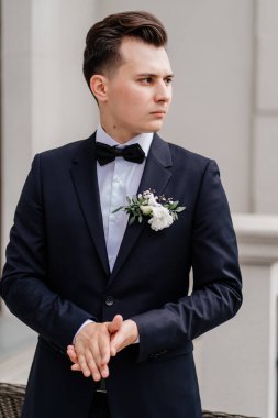 Siyah ceketli, çiçek tomurcuklu genç adam binanın yanındaki sokakta duruyor.