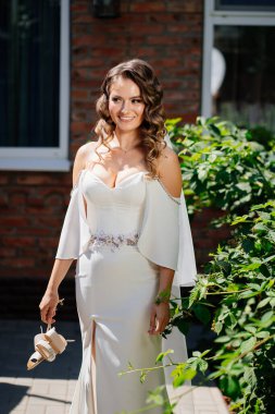 Uzun bukleli bir gelin, beyaz, zarif bir elbise ve elinde topuklu sandaletler.