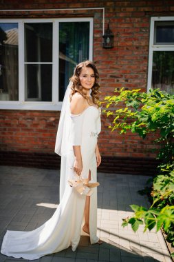 Uzun bukleli bir gelin, beyaz, zarif bir elbise ve elinde topuklu sandaletler.